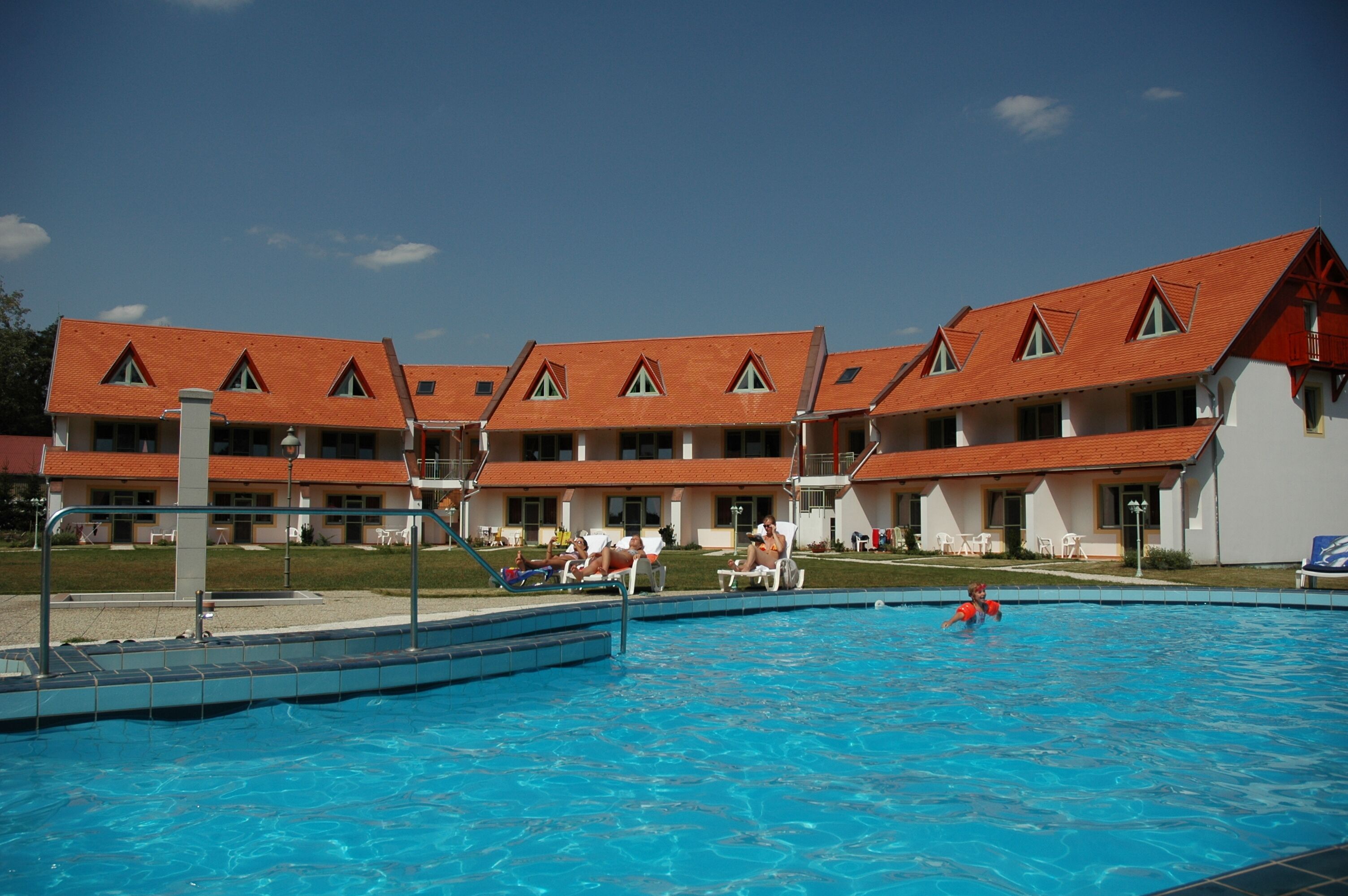 A swimming pool with a clear blue water surface, surrounded by a building with multiple balconies and a red roof.