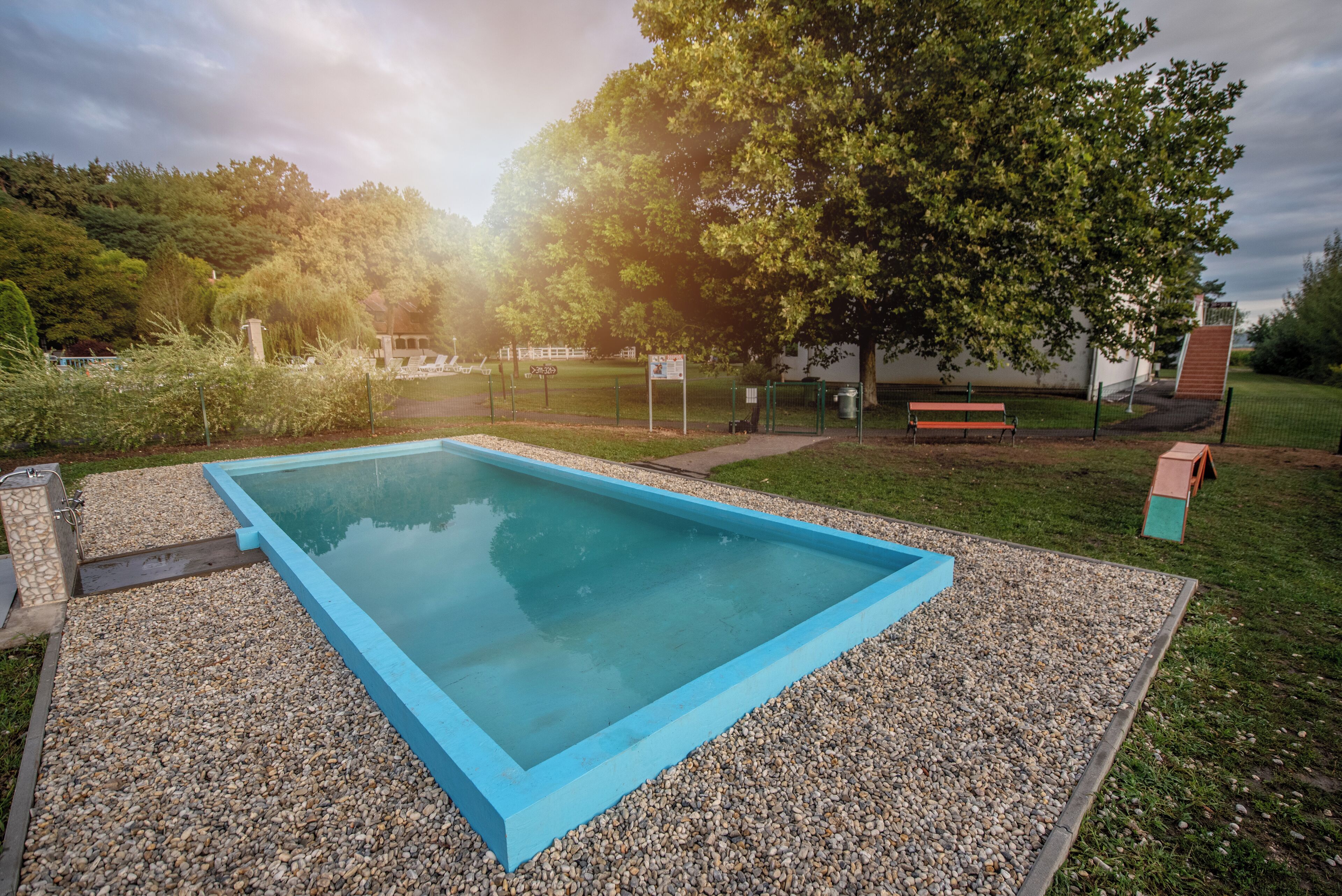 Una piscina rectangular con borde azul, rodeada de grava, con un banco y una pequeña construcción cerca.