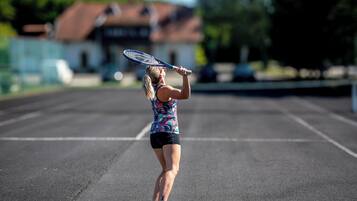 Een tennisser op een veld, met een huis op de achtergrond.