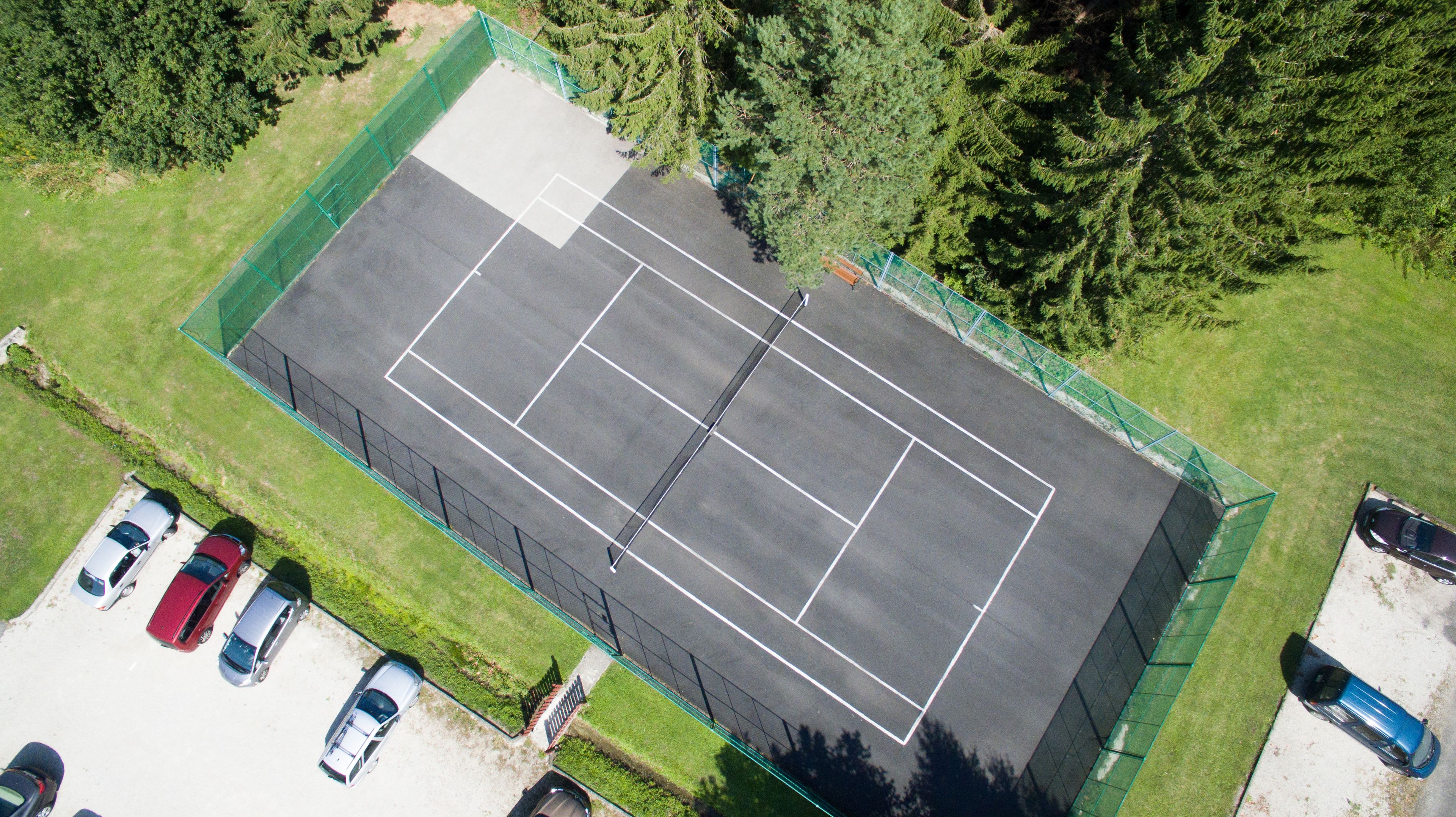 An aerial view of a tennis court surrounded by a green fence, with several cars parked nearby.