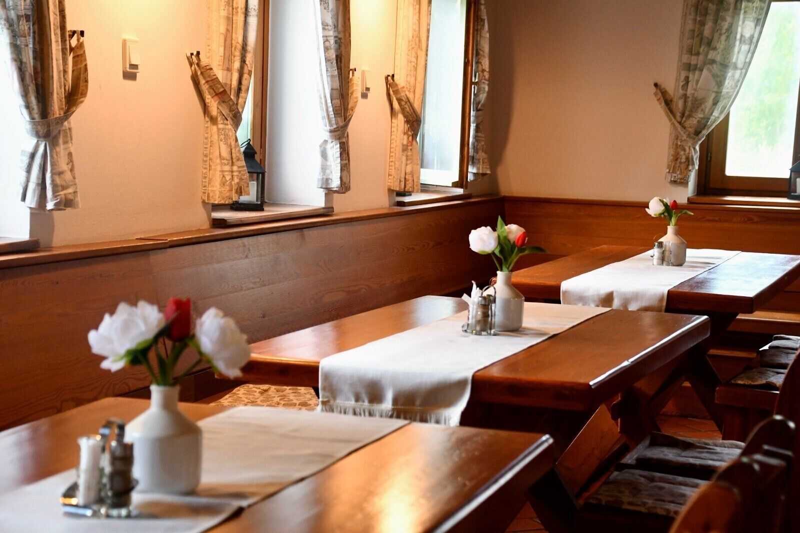 A dining area with wooden tables and benches, white tablecloths, and vases with flowers.