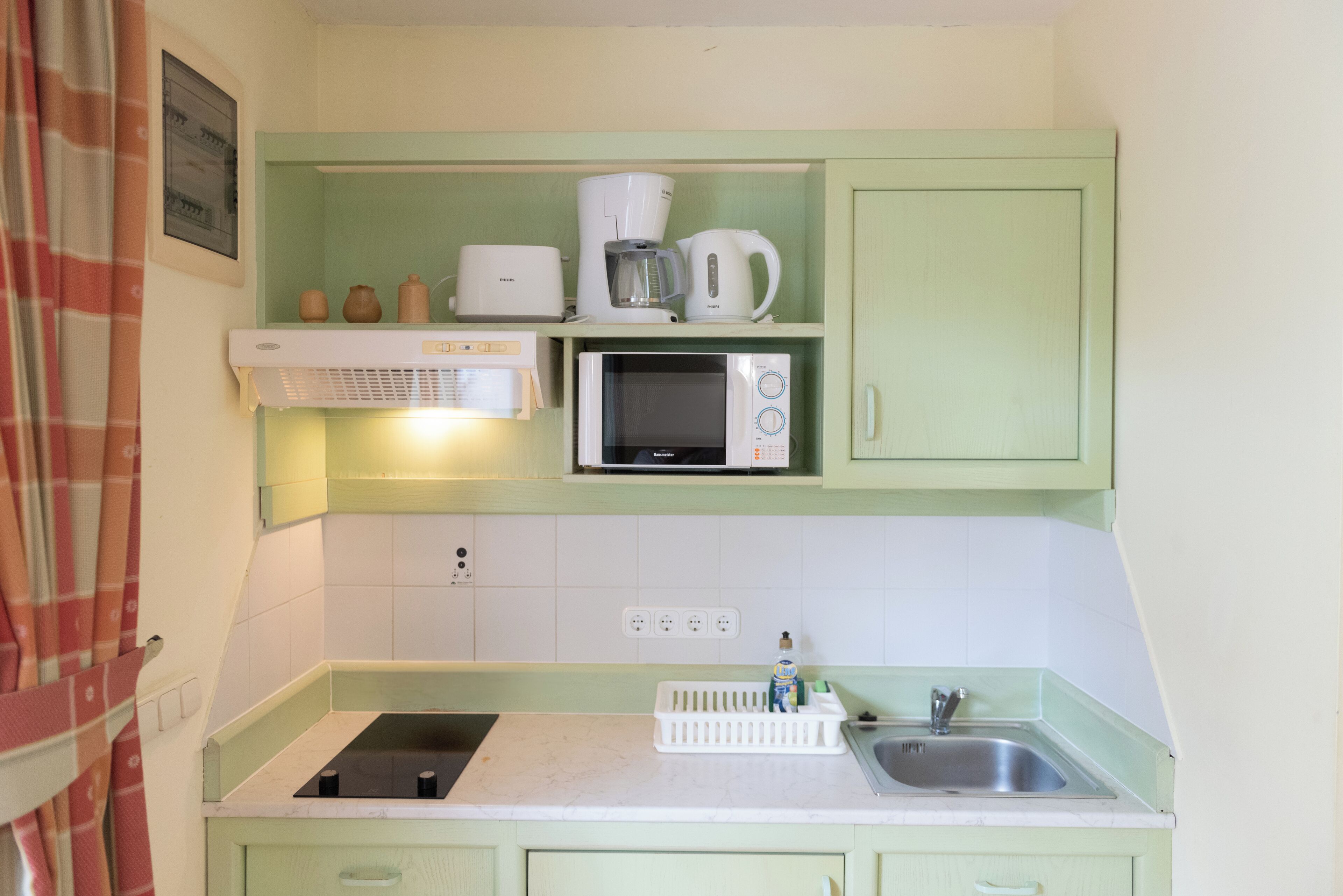 Cocina compacta con microondas, tostadora y cafetera sobre un mueble verde, un lavamanos y una pequeña encimera con un escurreplatos.