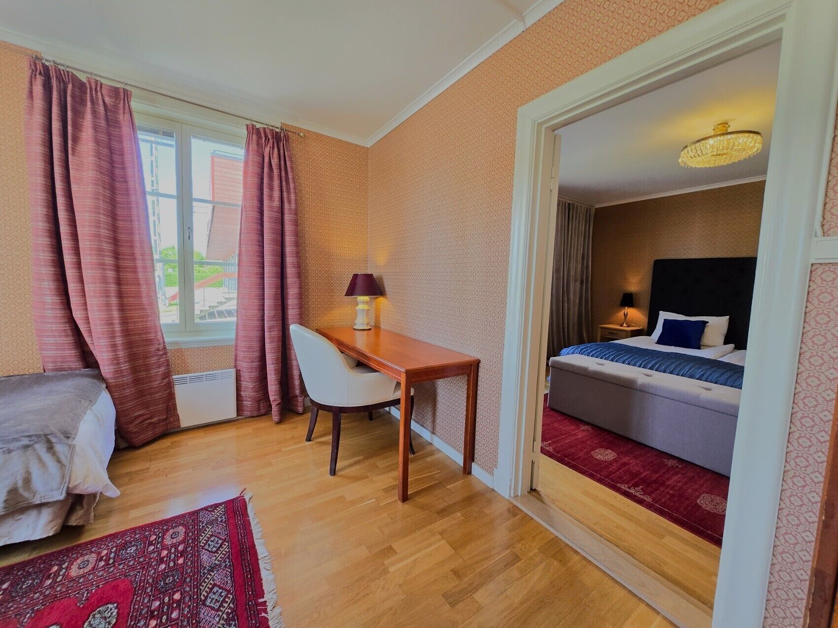 Une chambre d’hôtel avec un lit, un bureau et une chaise. Il y a une fenêtre avec des rideaux, un tapis rouge et un lustre.