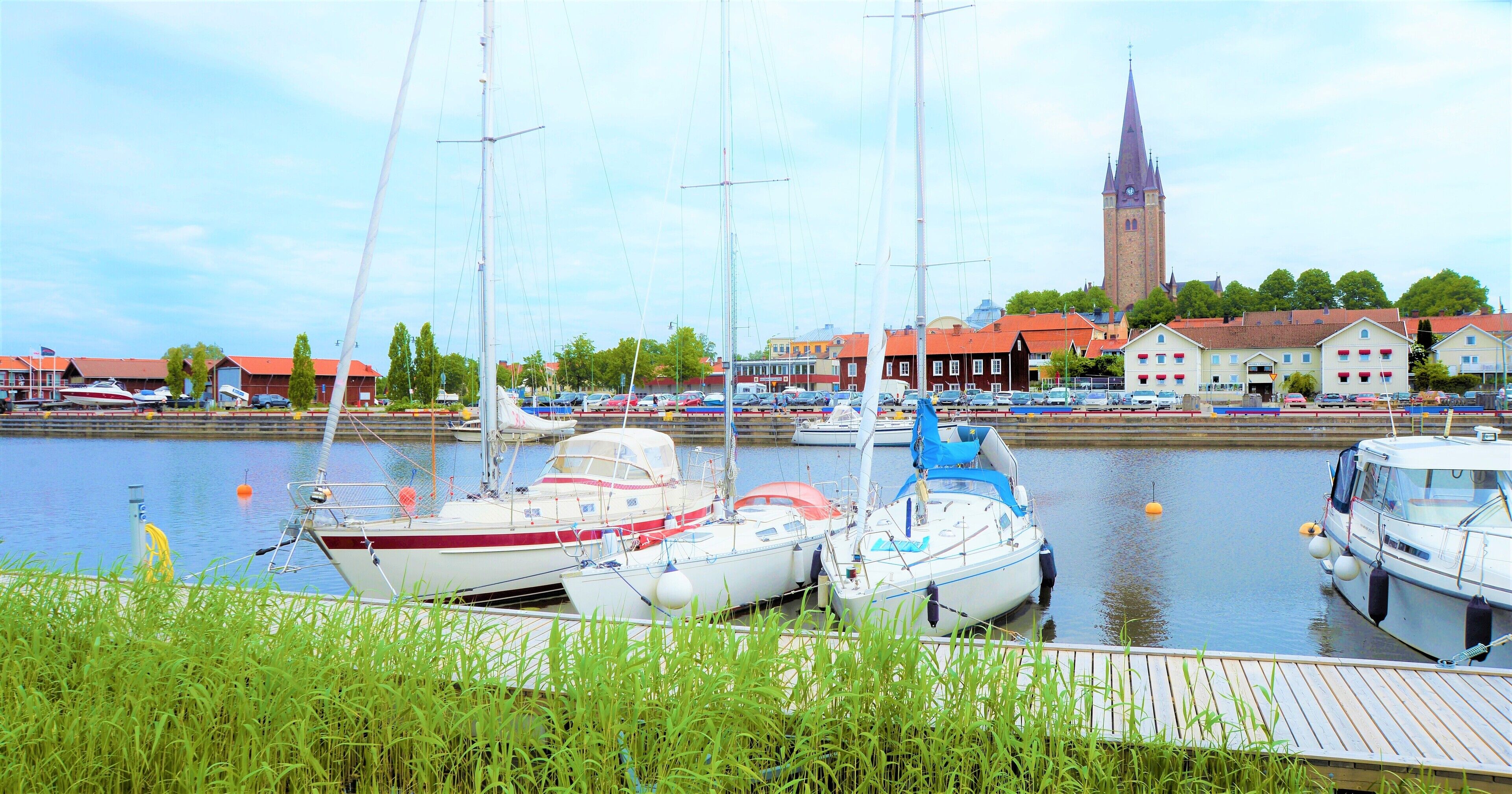 A marina with several boats docked, a church tower in the background, and a wooden dock in the foreground.