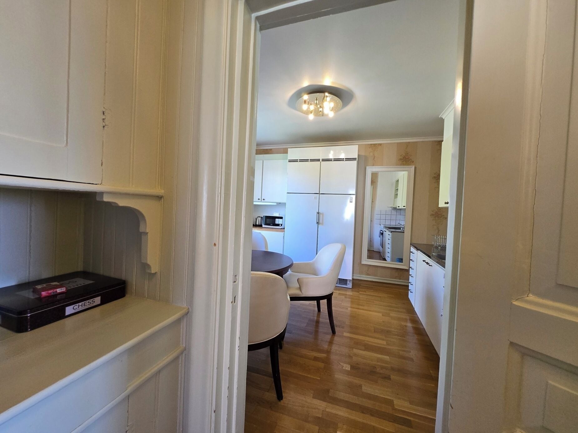 A narrow hallway leading to a dining area with a table and chairs, a kitchen with cabinets, and a ceiling light fixture.