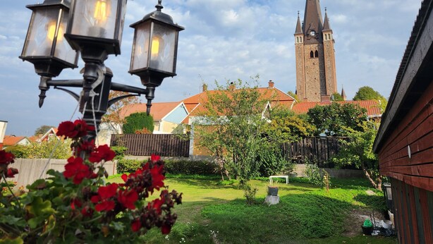 A church with a tall spire and a clock, surrounded by a well-maintained garden with red flowers and a wooden fence. - Vänerport Lakefront (Mariestad)