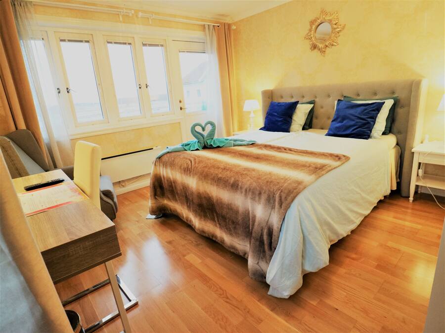 A hotel room with a bed, bedside table, lamp, and a view of the outdoors through the window.