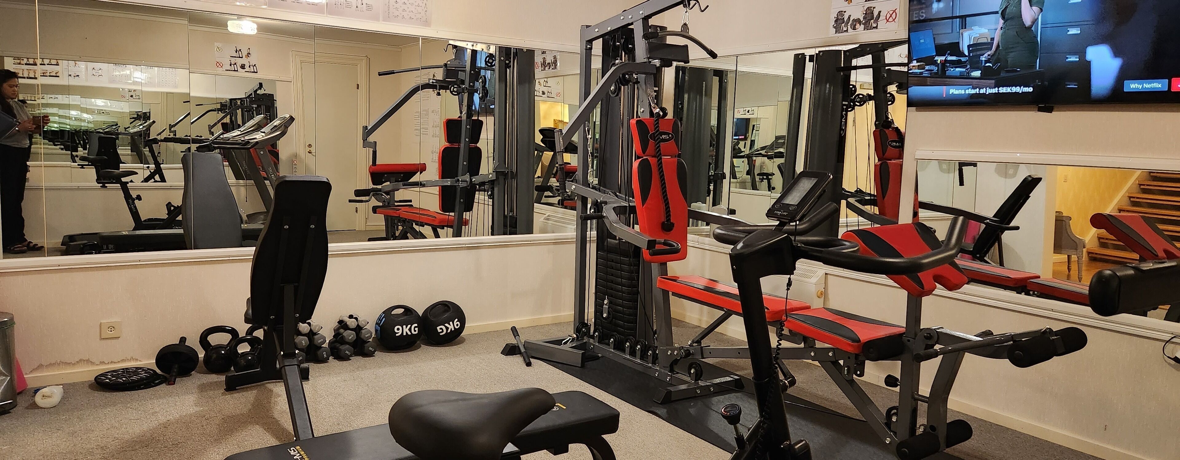 A gym with various exercise equipment, including a bench press, dumbbells, and a TV mounted on the wall.