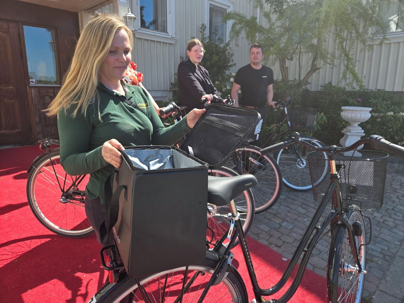 A woman holding a bicycle with a basket, standing on a red carpet outside a building with a door and windows.