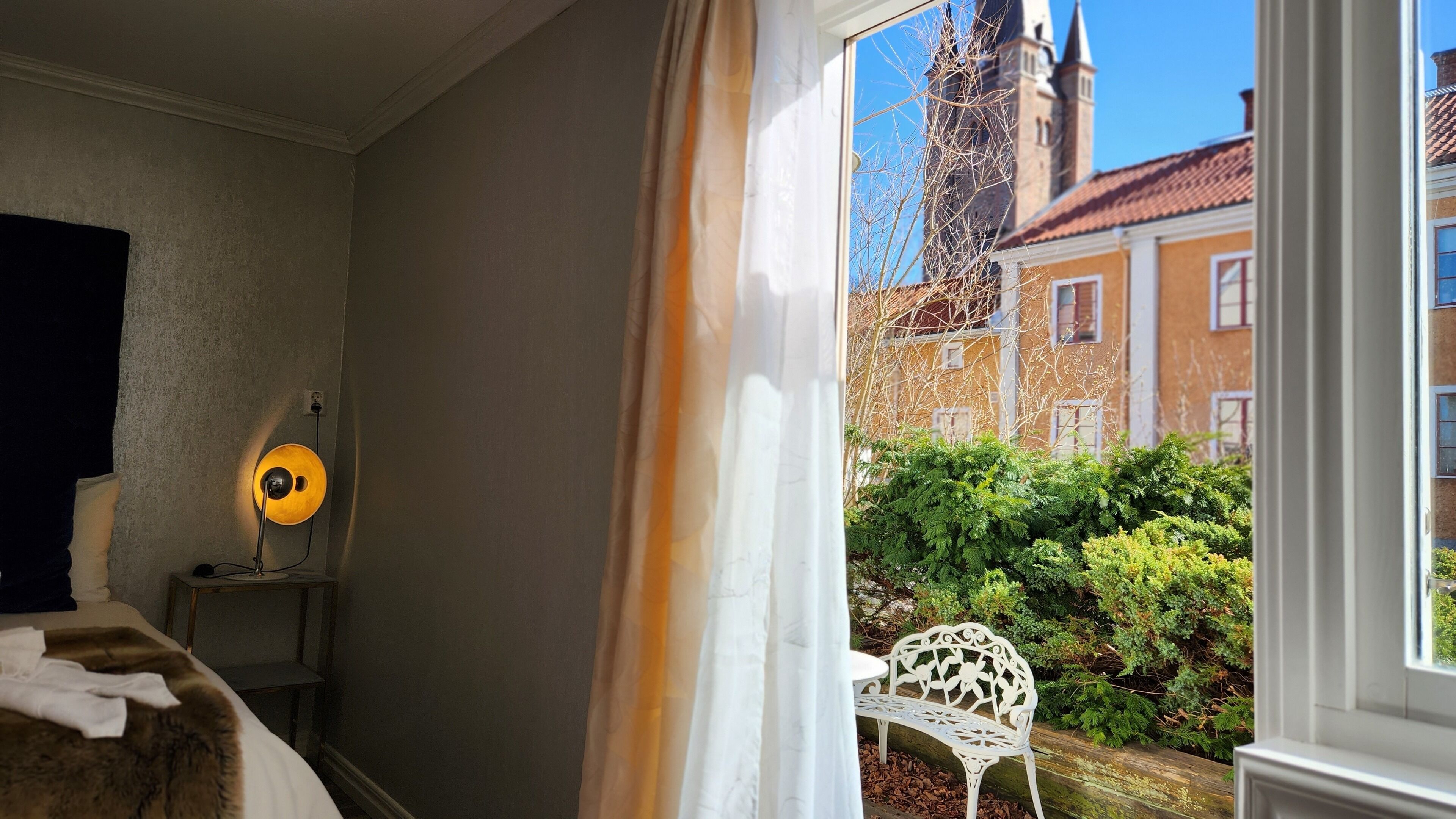 Una habitación con una cama, una lámpara, una silla y una ventana con vistas a un edificio y zonas verdes.