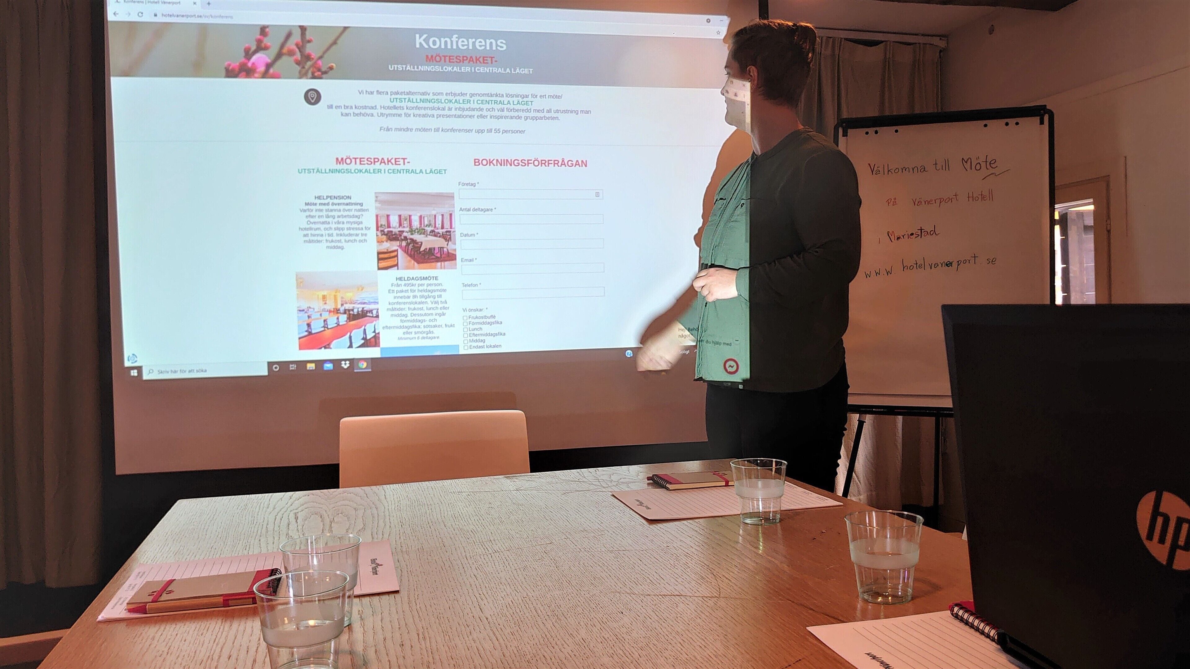 En presentation i ett konferensrum med en skärm som visar information om hotellet, en whiteboard med handskrivna anteckningar och ett bord med dokument och glas.