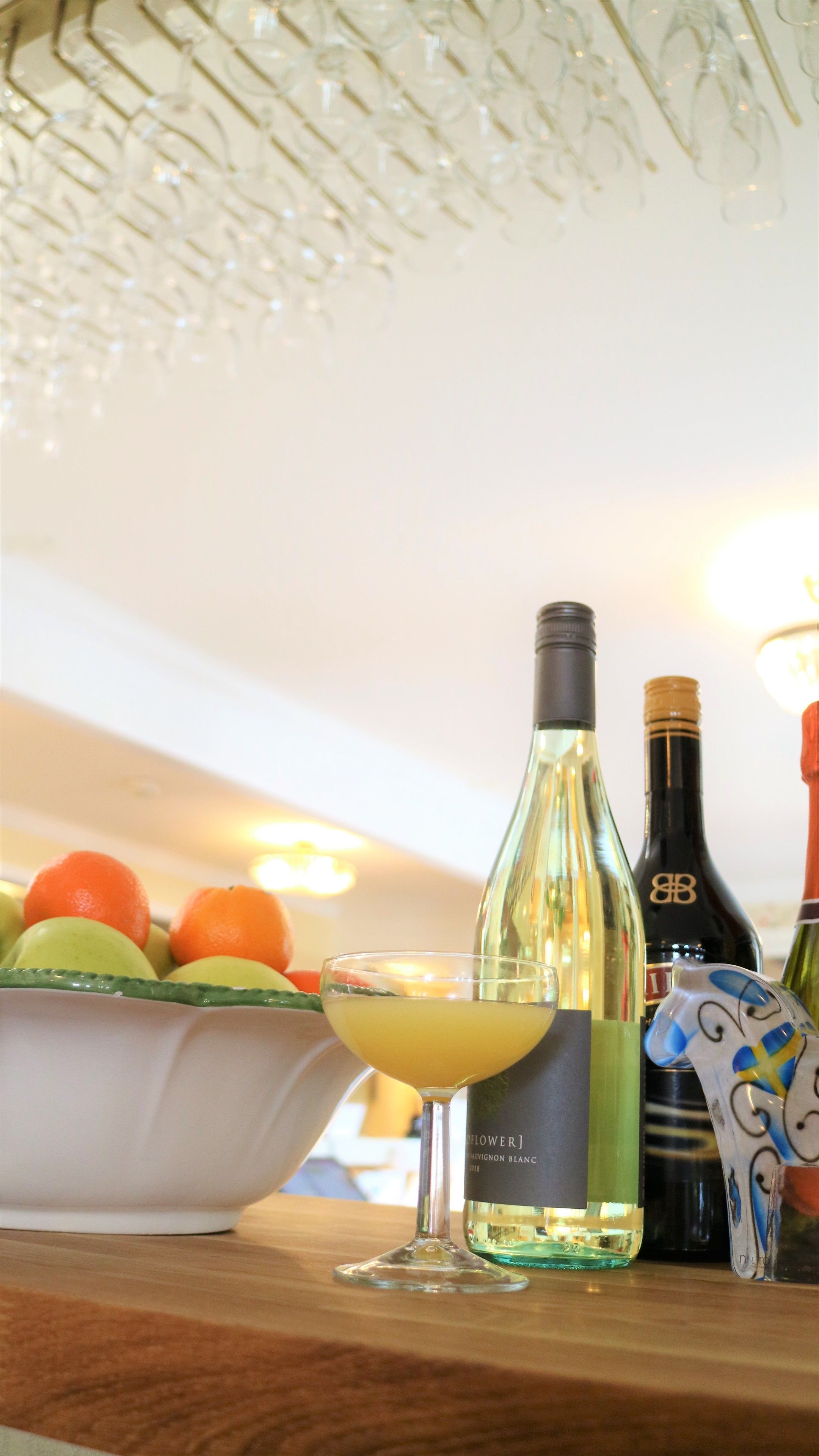 A bar setting with a glass of white wine, a bottle of wine, and a bowl of fruit.