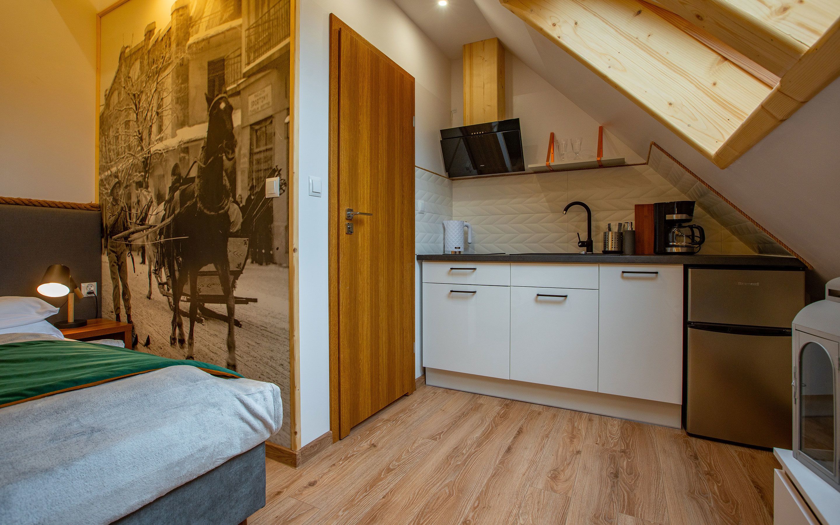 A compact bedroom with a bed, a wooden door, and a wall-mounted picture. Adjacent is a kitchenette with a stove, sink, and storage cabinets.