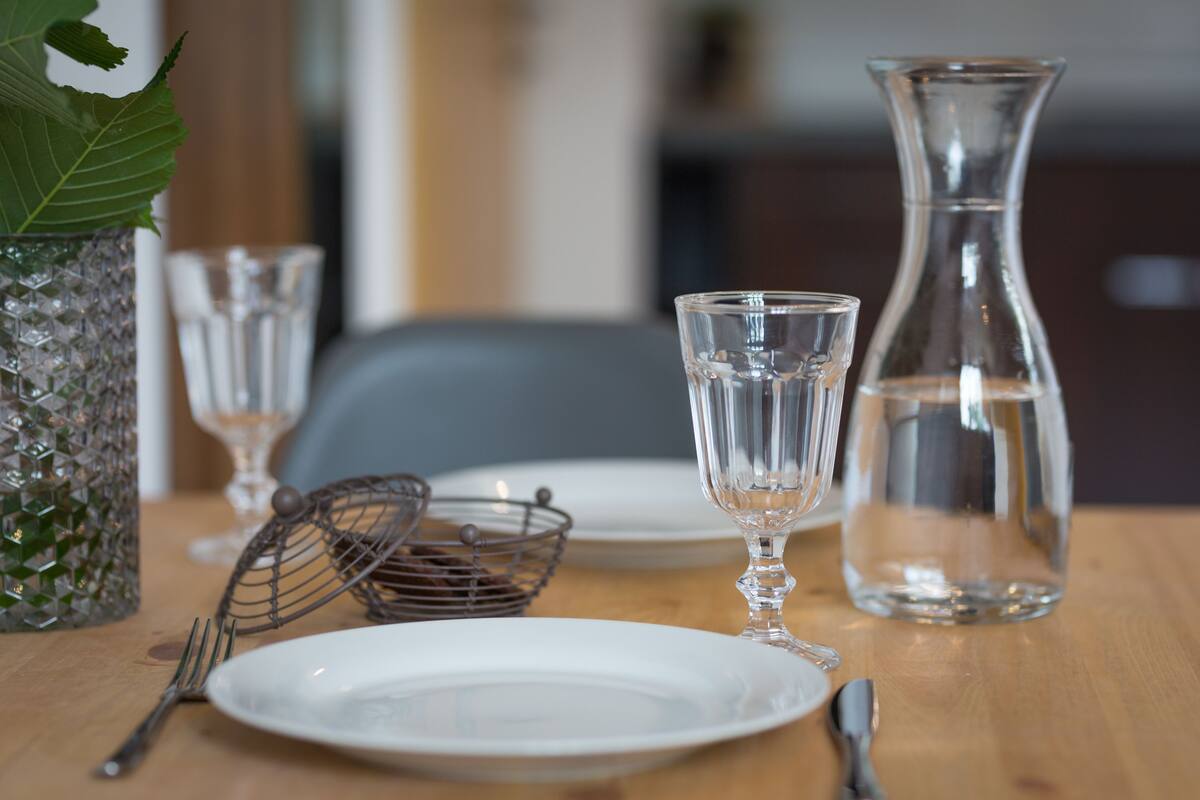 Une table à manger avec une carafe en verre transparent, deux verres à vin, une passoire en métal, une fourchette, une cuillère et une assiette blanche.