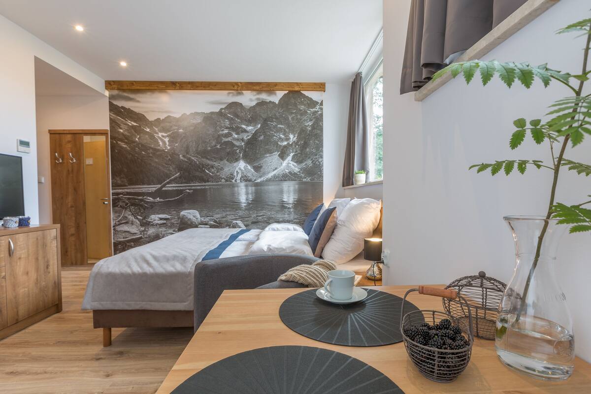 Une chambre à coucher avec un lit, une table en bois, une plante, un tableau et une fenêtre.