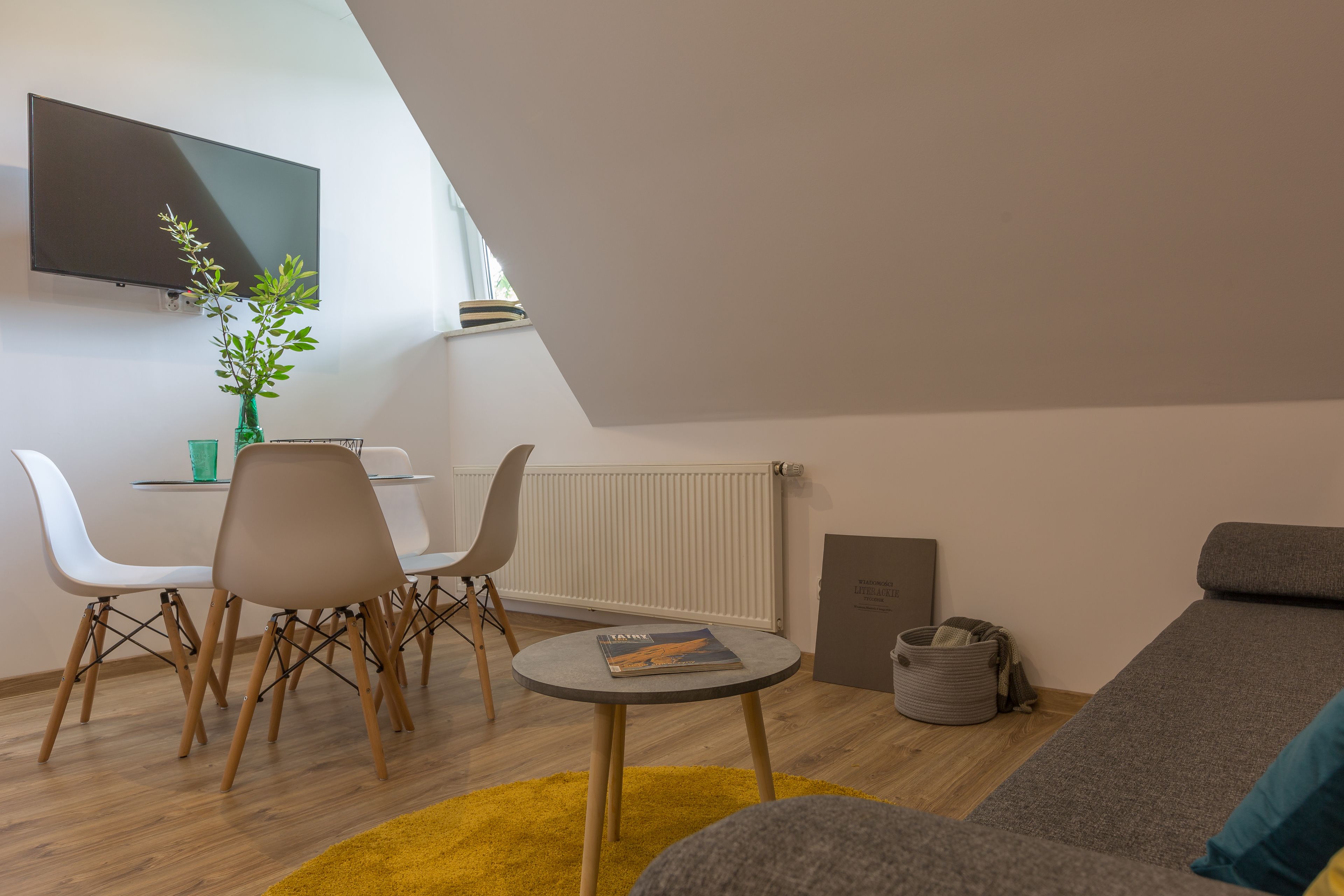 A modern living room with a sofa, a dining table with chairs, a small round table with a magazine, a plant, and a TV mounted on the wall.