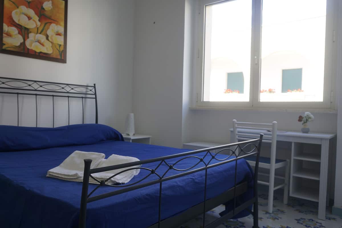 Une chambre à coucher avec un lit, une table de chevet, une chaise, une fenêtre et un tableau au mur.