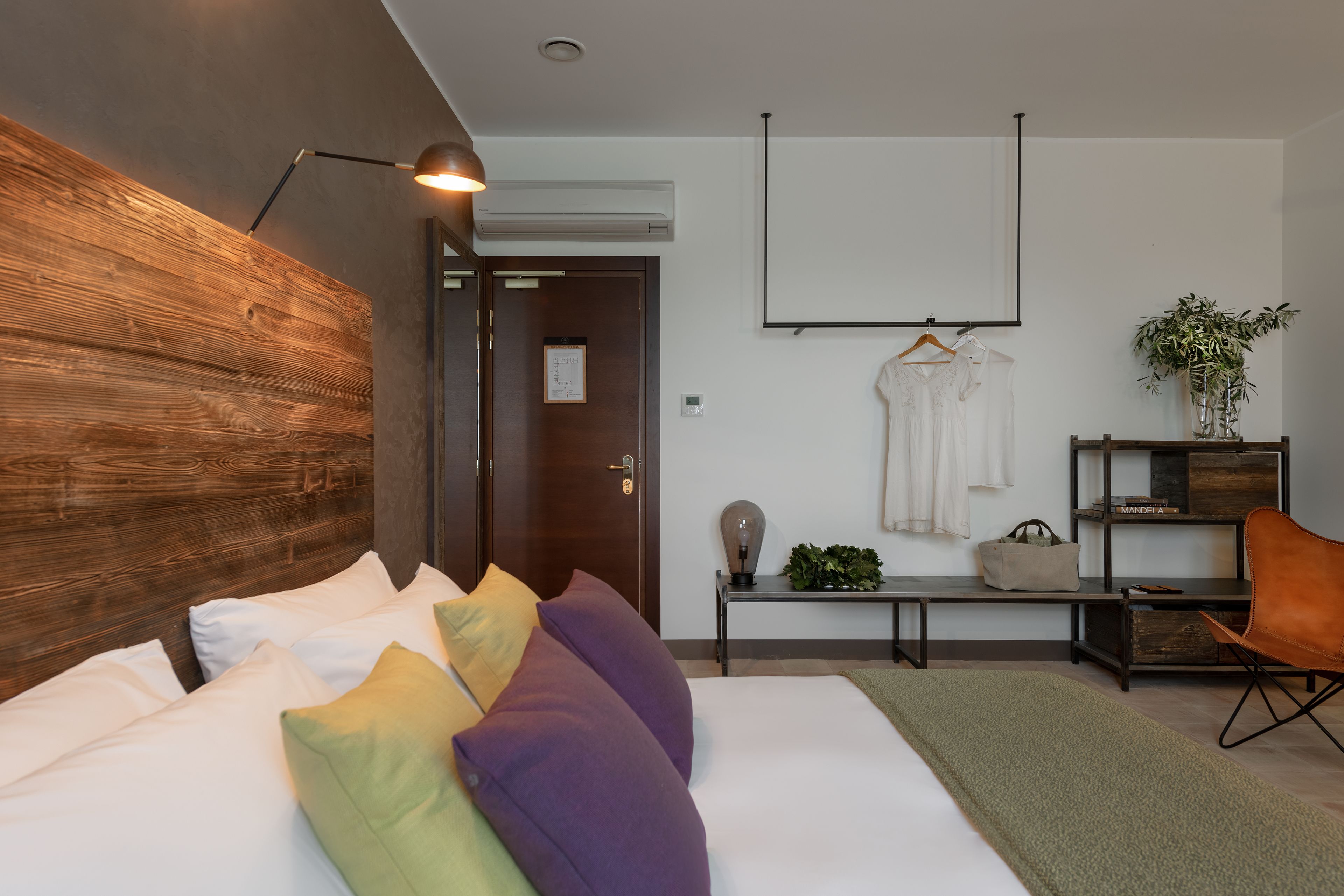 Una habitación de hotel moderna con cabecera de madera, cama con almohadas blancas y coloridas, escritorio con lámpara, perchero y planta en maceta.