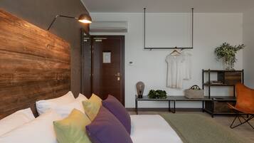 Una habitación de hotel moderna con cabecera de madera, cama con almohadas blancas y coloridas, escritorio con lámpara, perchero y planta en maceta.