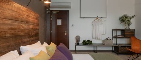 A modern hotel room with a wooden headboard, a bed with white and colorful pillows, a desk with a lamp, a hanging coat rack, and a potted plant.