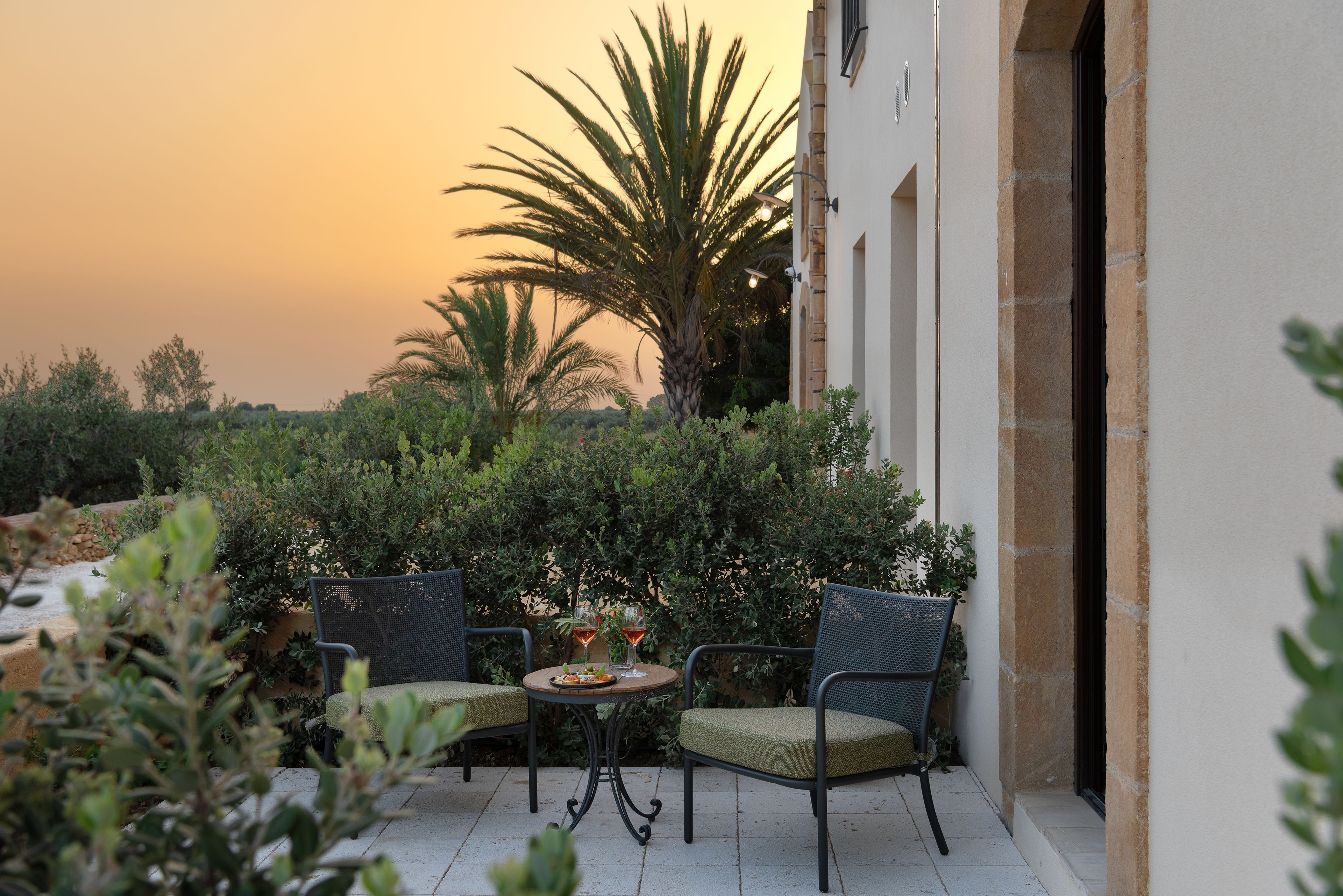 An outdoor patio with two wicker chairs and a small round table, surrounded by lush greenery and a palm tree.