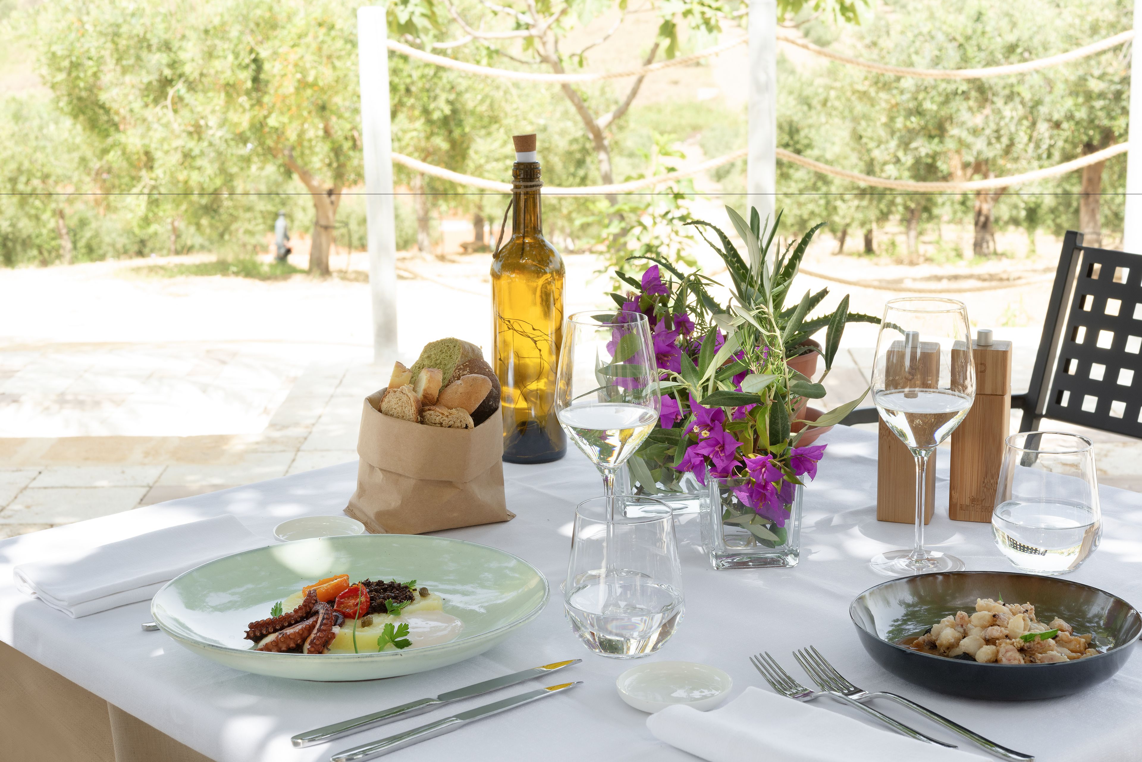 Mesa al aire libre para dos personas, con platos de comida, copas de vino y una botella de aceite de oliva.