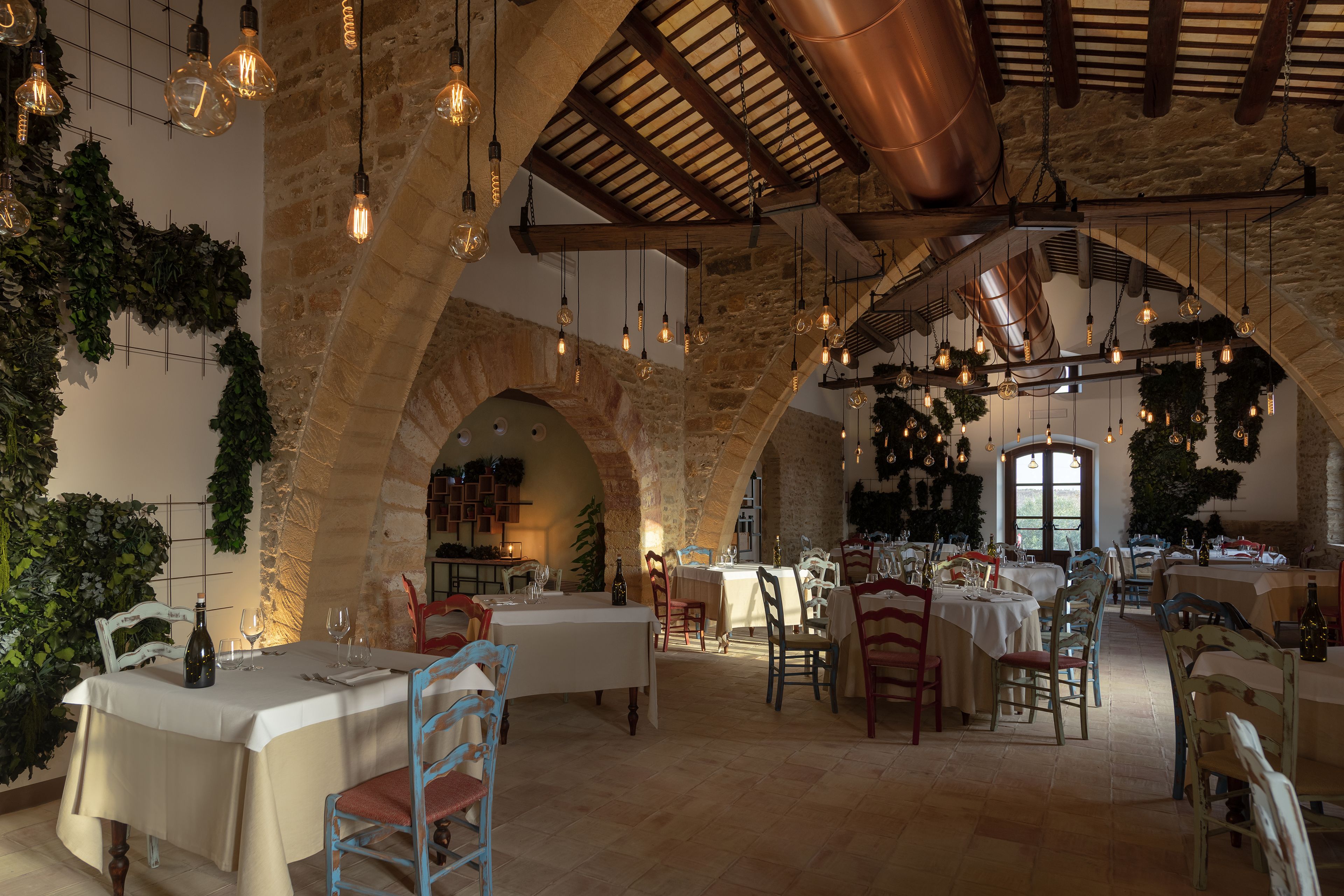 Een restaurant met rustieke stenen bogen, hangende lampen en houten balken. Tafels zijn gedekt met witte tafelkleden en stoelen.