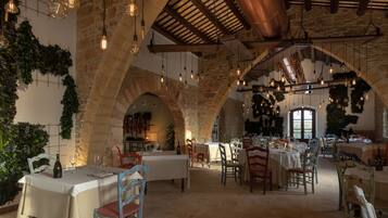 Een restaurant met rustieke stenen bogen, hangende lampen en houten balken. Tafels zijn gedekt met witte tafelkleden en stoelen.
