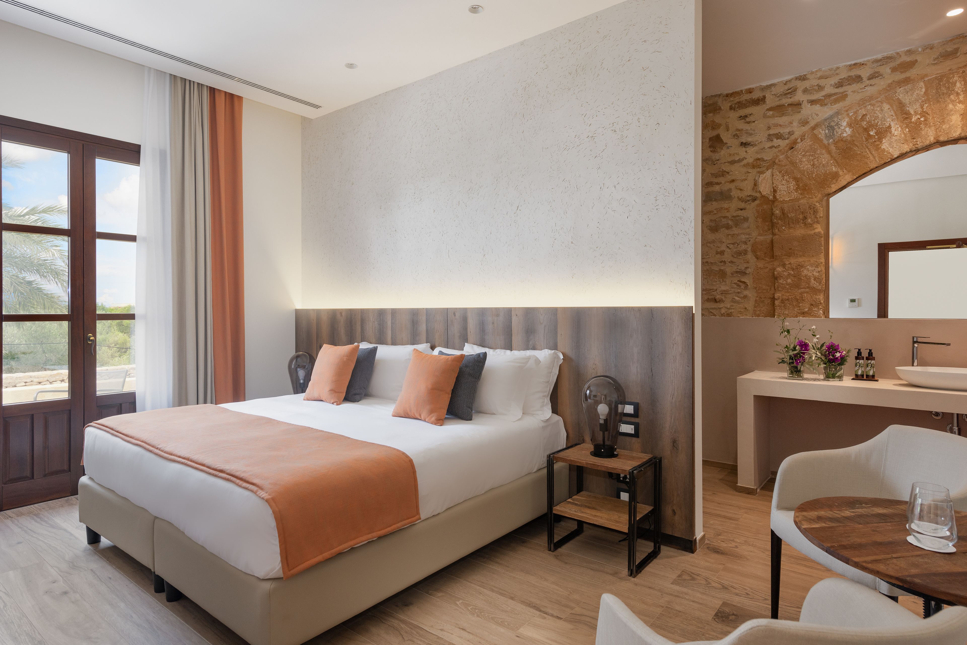 Ein modernes Hotelzimmer mit einem großen Bett, einem hölzernen Nachttisch, einem kleinen Tisch mit Stuhl und einer Wand aus Stein.
