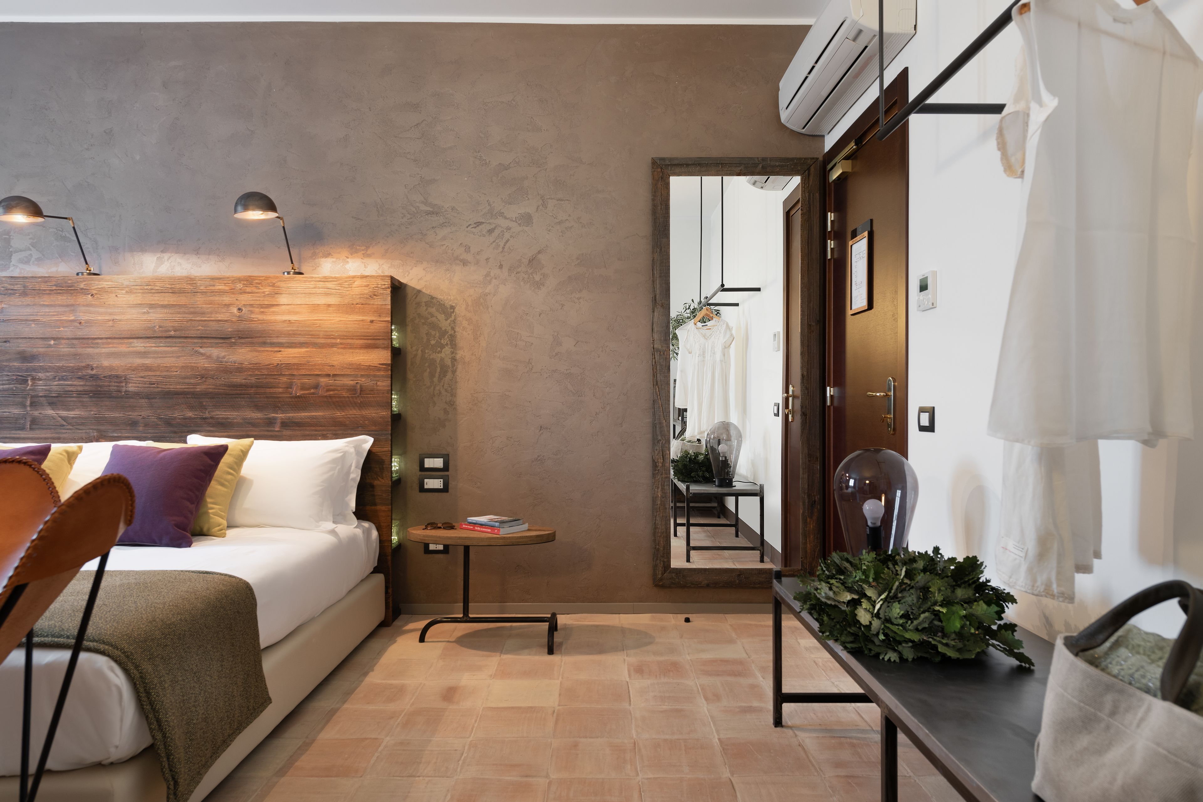 Een moderne hotelkamer met een houten hoofdbord, een tafeltje met boeken, een stoel en een spiegel die de kamer weerspiegelt.