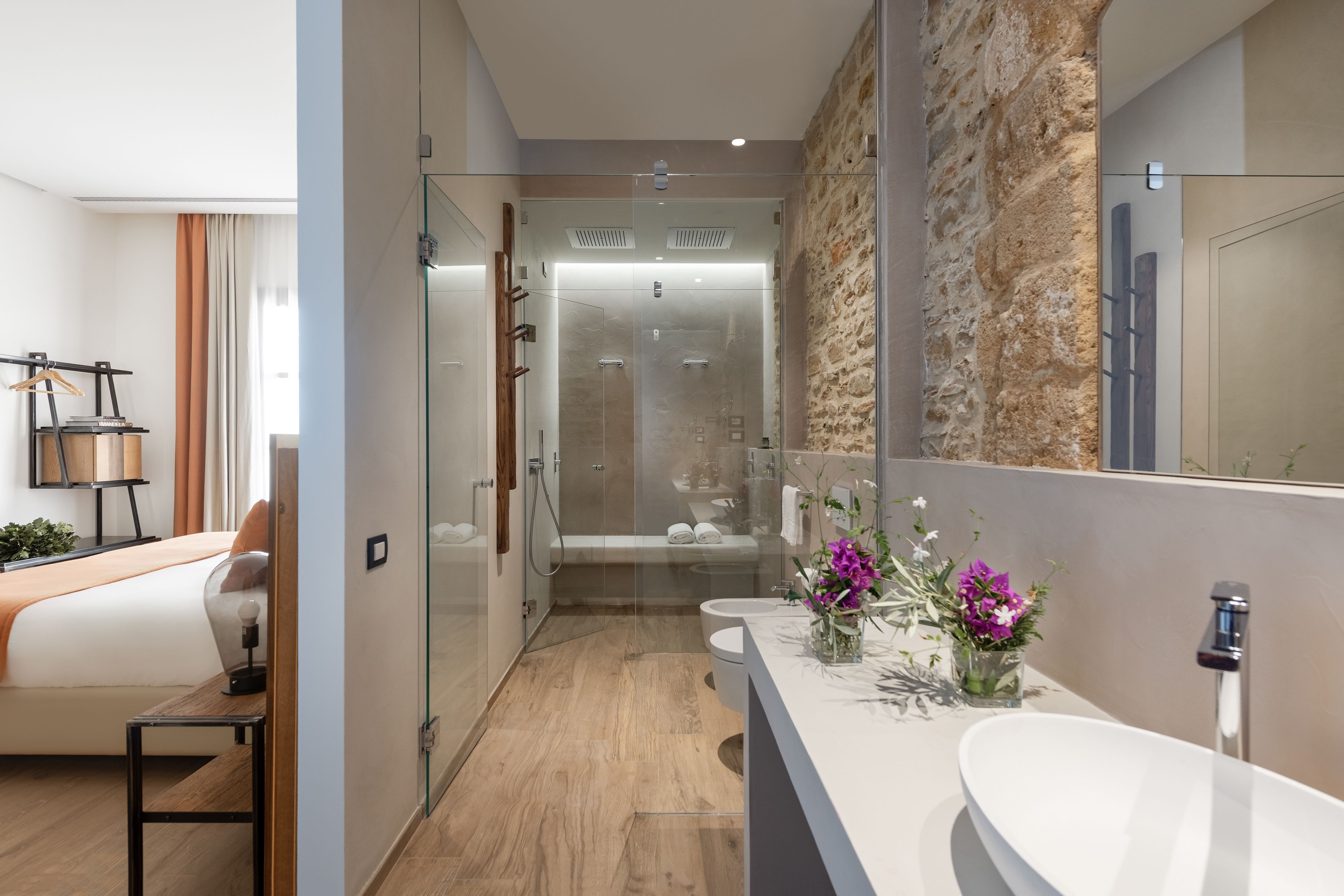 A modern bathroom with a glass shower enclosure, a white sink with a vase of flowers, and a wooden floor.