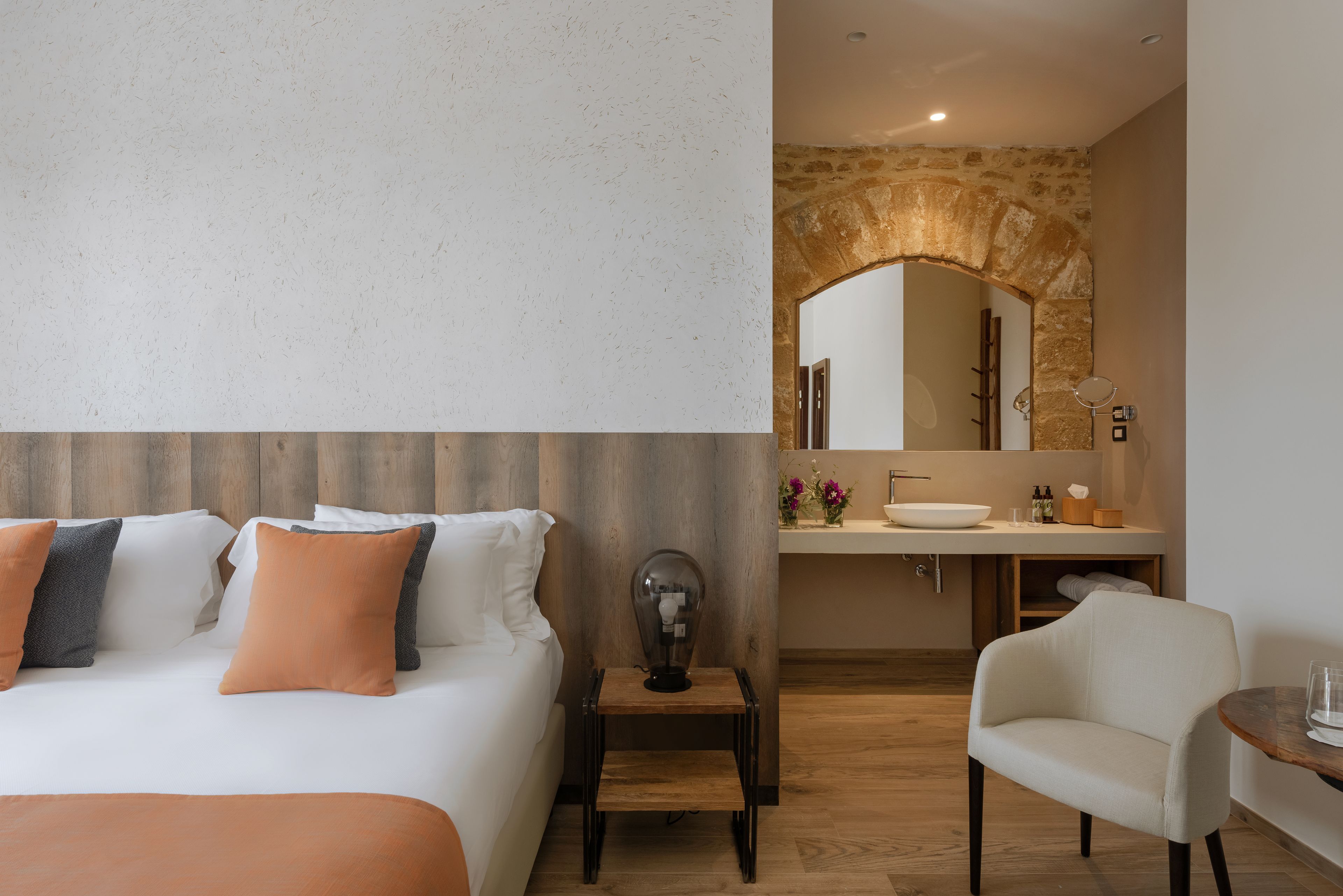 A hotel room with a bed, a nightstand, a chair, a bathroom with a sink and mirror, and a stone wall.