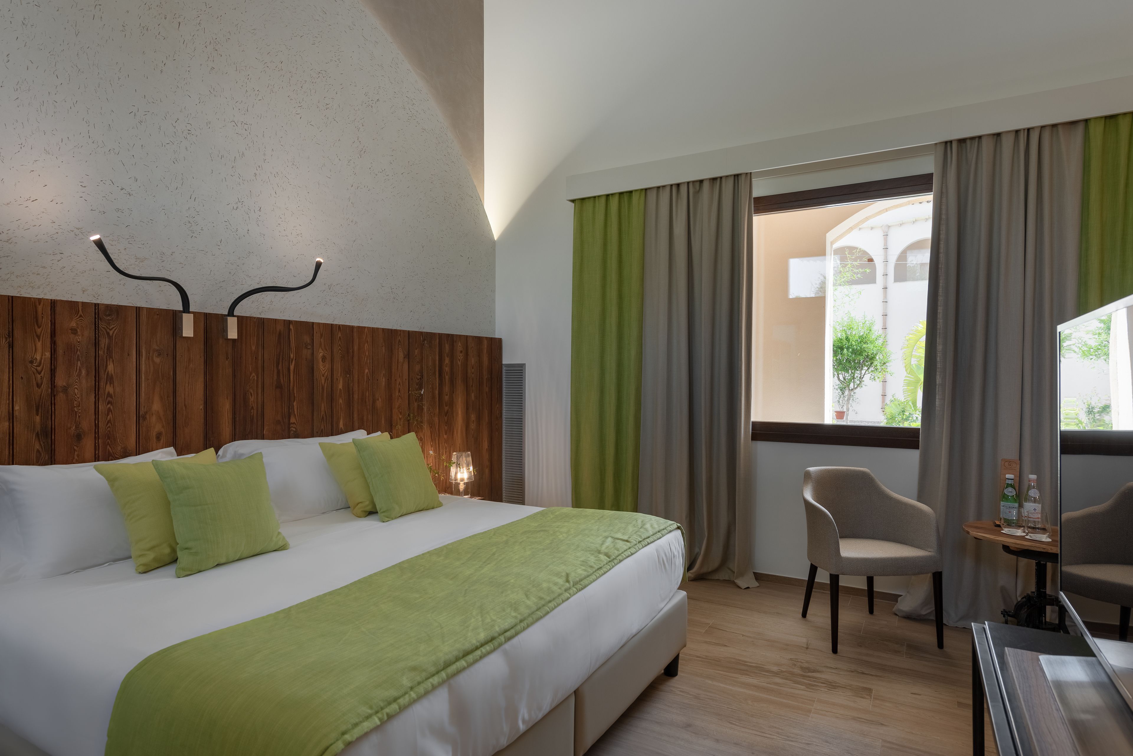 A hotel room with a large bed, a wooden headboard, a green throw blanket, a chair, a small table with a bottle, and a view through a window with curtains.