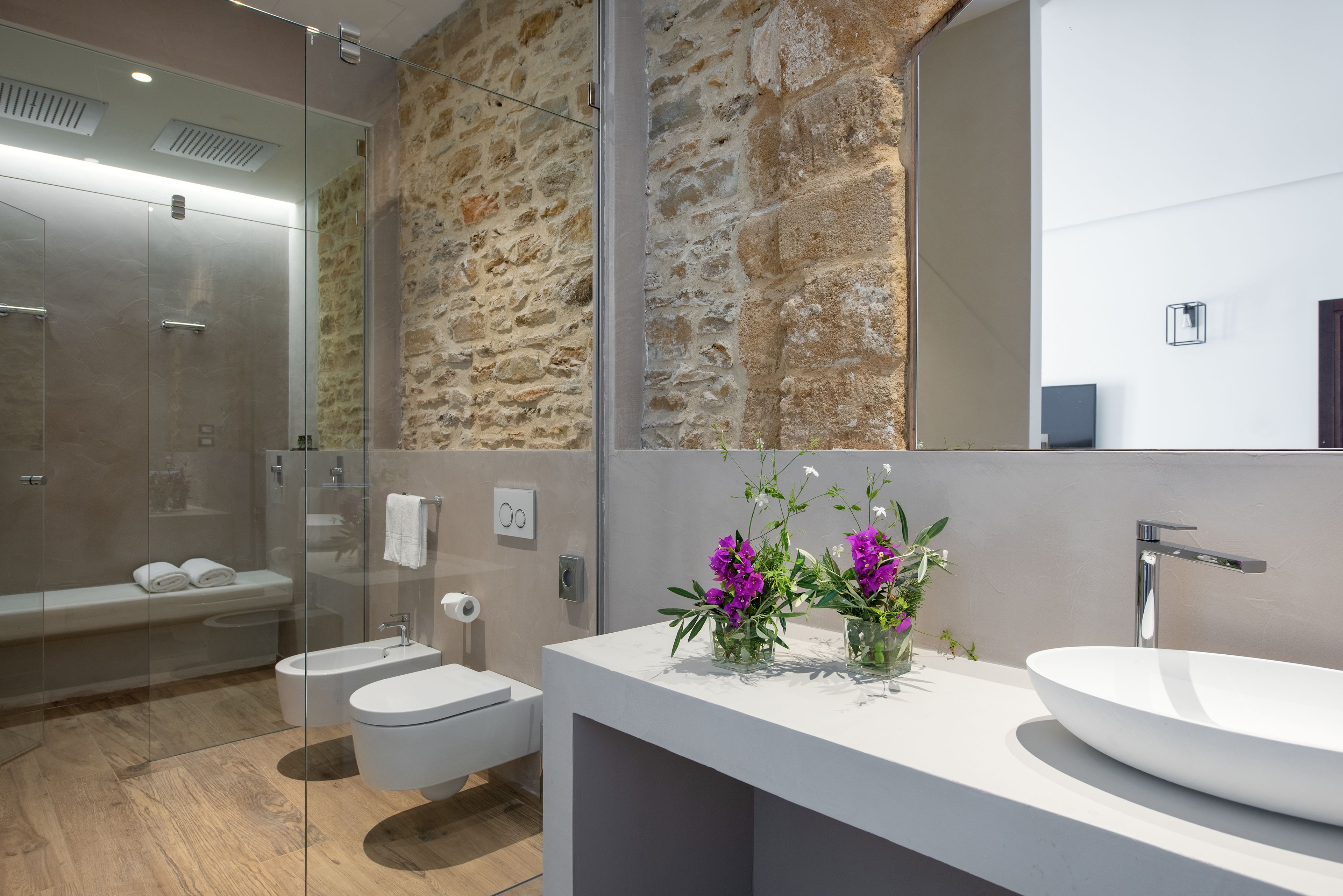 Een moderne badkamer met een stenen muur, een douche met glazen wanden, een wit wastafelmeubel en een toilet.