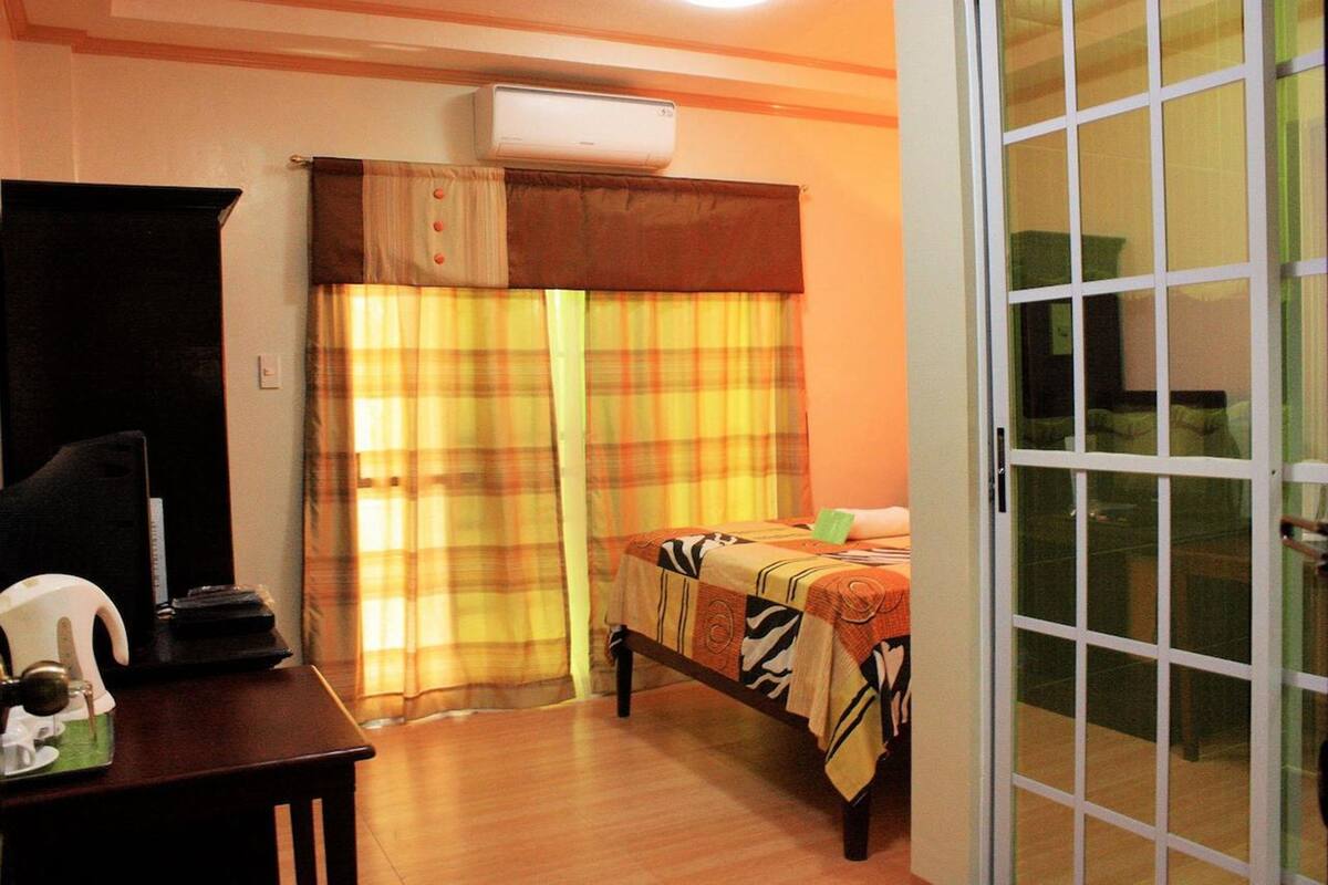 Une chambre avec un lit, un bureau et une chaise, un climatiseur et une fenêtre avec des rideaux.
