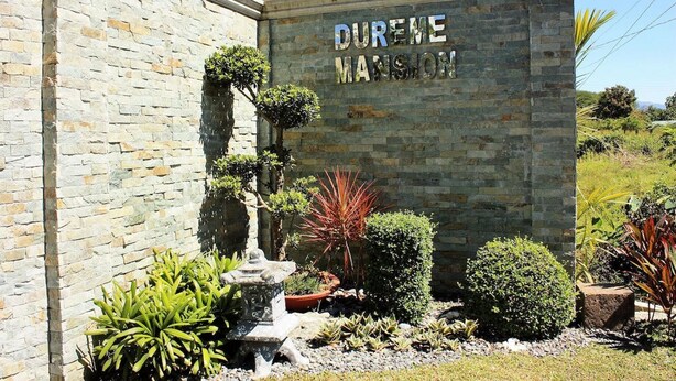 Un mur de pierre sur lequel est inscrit le texte « DURENEM MANSON », ainsi qu’un jardin agrémenté de diverses plantes et d’une fontaine en pierre.