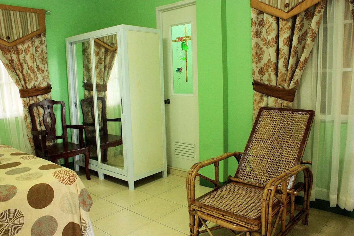 Une pièce avec un mur vert, une armoire, un lit recouvert d’une couvre-lit à pois et deux fauteuils en osier.