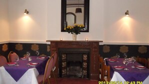A dining area with two tables set for four, each with a purple tablecloth. There are candles on the mantelpiece above the fireplace, and a vase with yellow flowers on the table. - Thorpe Lodge Hotel (Peterborough)