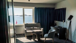 Habitación de hotel con sofá, sillón y una pequeña mesa. Un ventanal grande ofrece vistas al mar y una bandera.