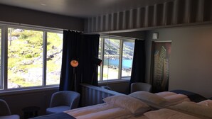 Habitación de hotel con cama, dos sillas, una lámpara y vistas a un lago y montañas a través de amplios ventanales.