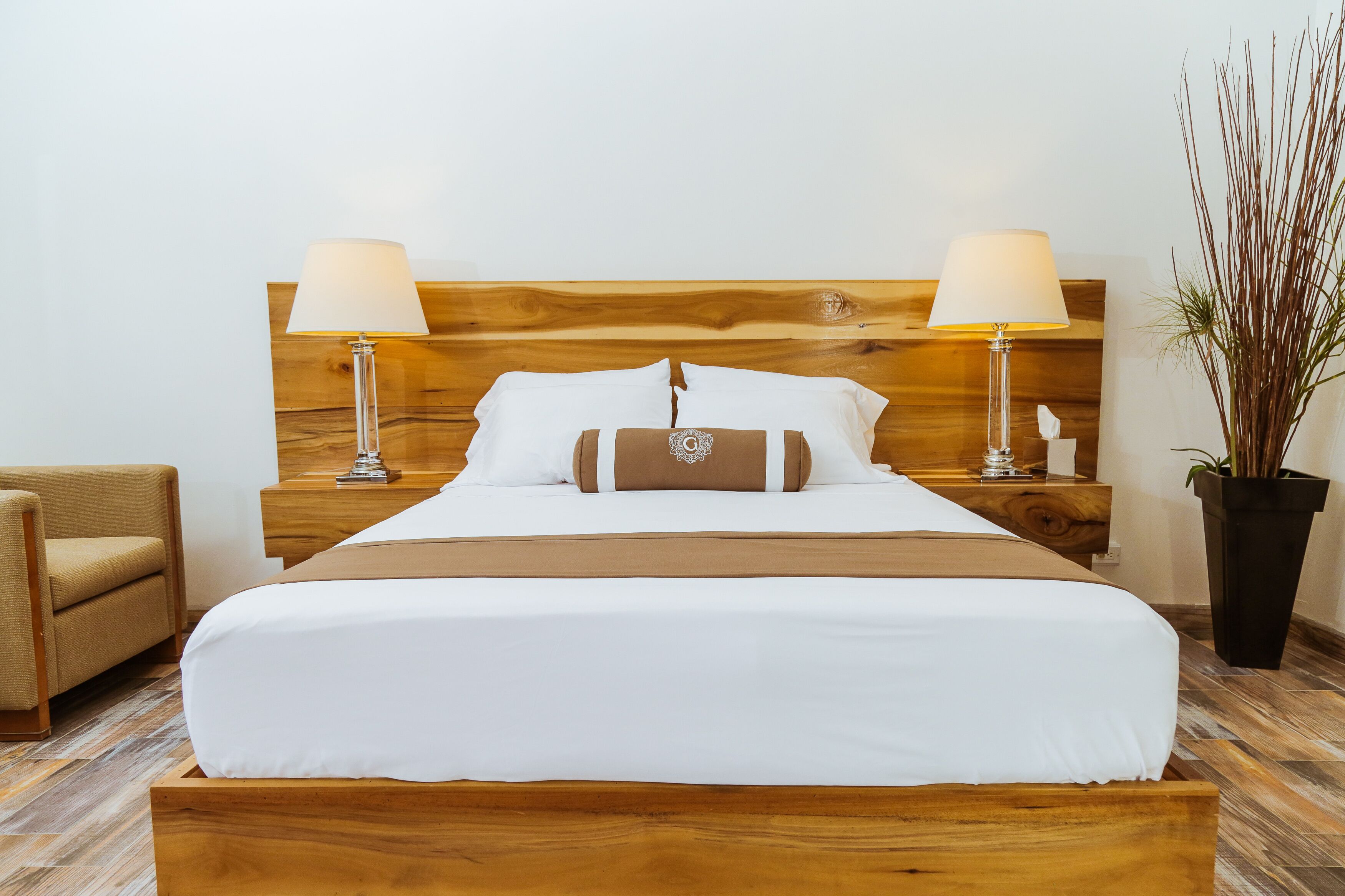 A neatly made bed with a wooden headboard, two bedside lamps, a bedside table with a lamp, a potted plant, and a chair.