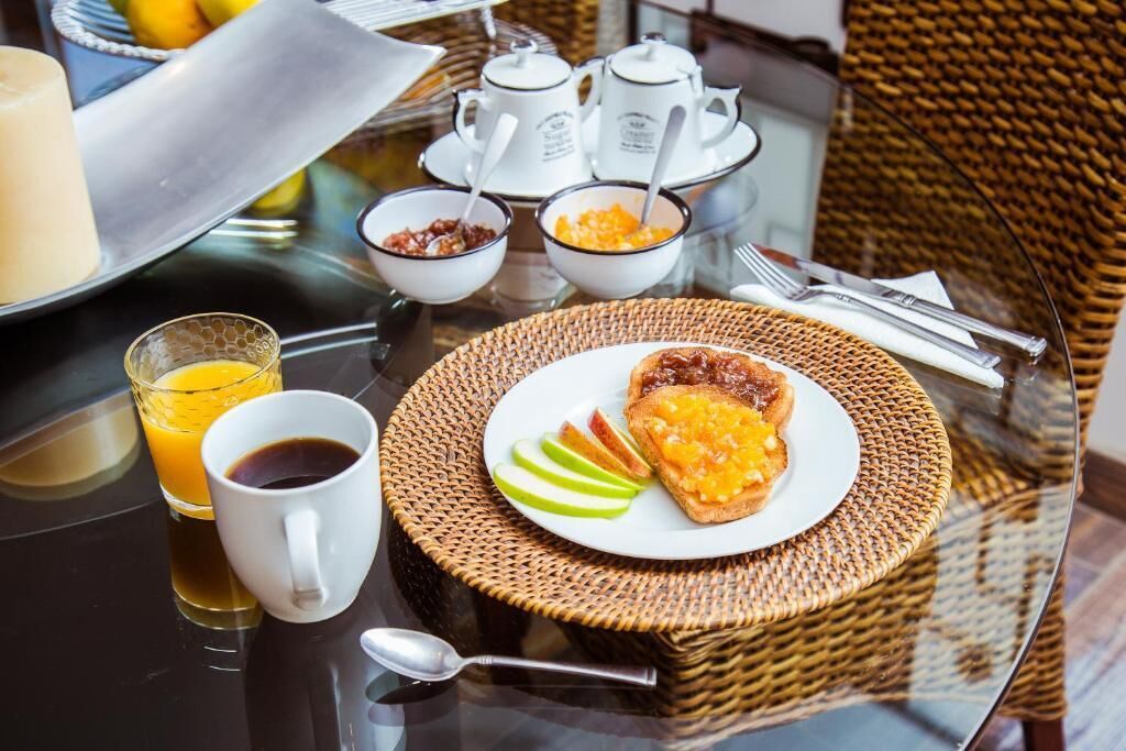 Une table en verre dressée pour le petit-déjeuner, avec des tartines, des pommes et du jus d’orange, accompagnée de café et de thé.