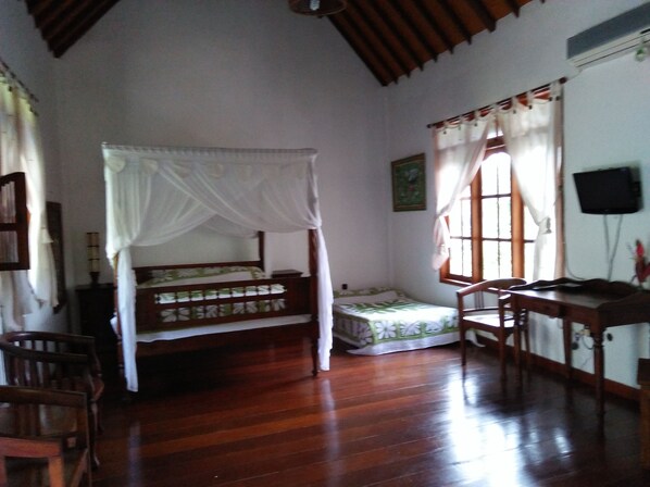 A room with a wooden floor, a bed with a canopy, a desk, chairs, and a television on the wall.