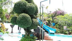 A pool area with a blue slide, surrounded by well-maintained greenery and a stone wall. - Puri Eling Blimbingsari (Melaya)