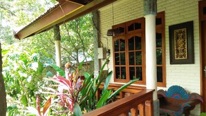 A wooden porch with a bench, a window with a decorative frame, and lush greenery. - Puri Eling Blimbingsari (Melaya)