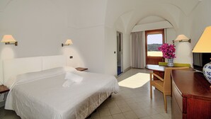 A hotel room with a large bed, two bedside lamps, a desk with a chair, a vase with flowers, and a view of the outdoors through a door. - Mursia Resort & Spa (Pantelleria)