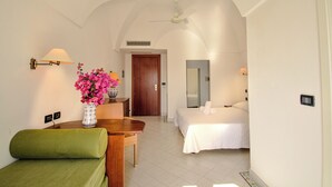 A hotel room with a bed, a green sofa, a wooden table with a vase of pink flowers, a ceiling fan, and a door leading to another room. - Mursia Resort & Spa (Pantelleria)