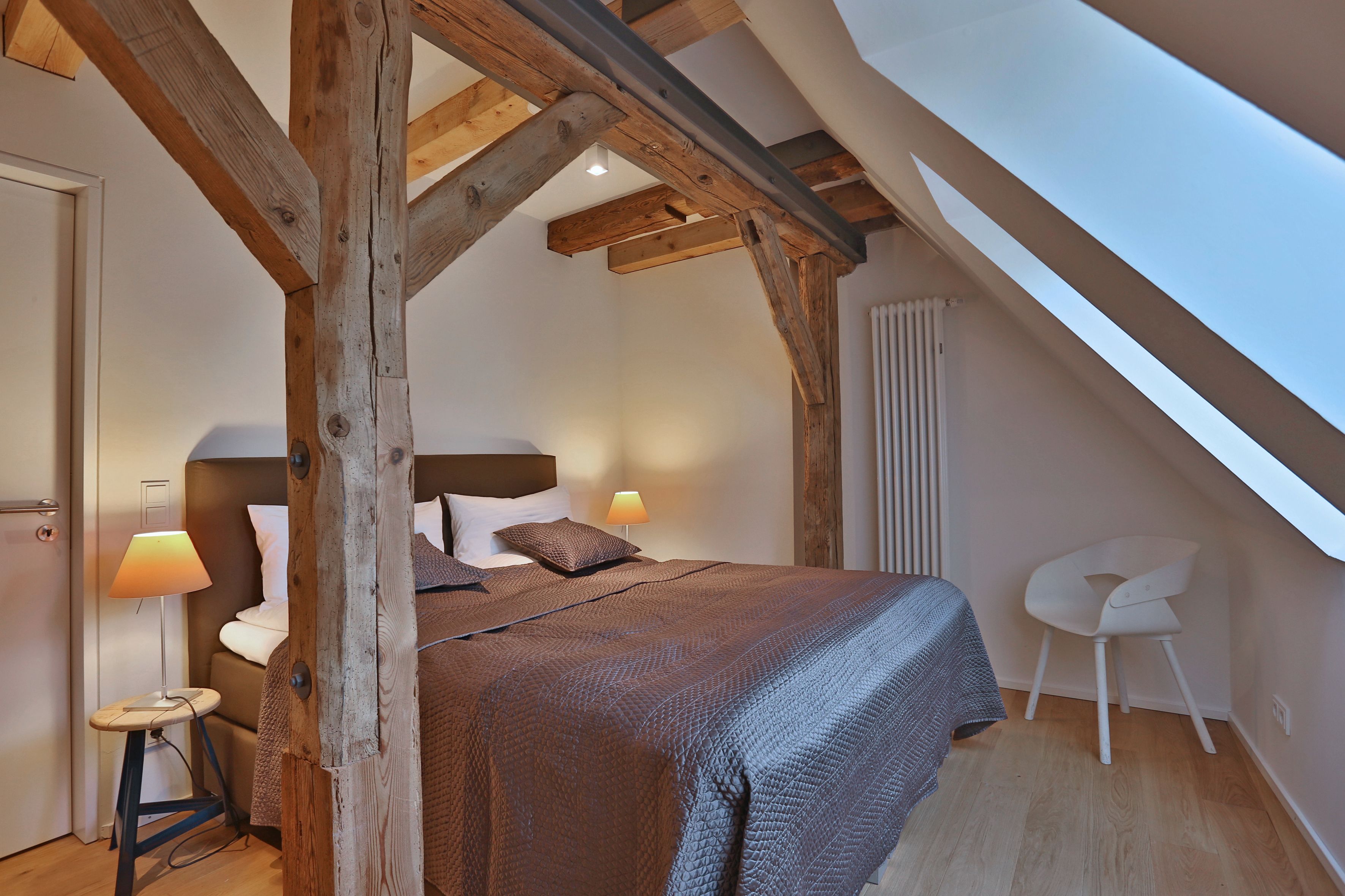 Un dormitorio con techo inclinado, vigas de madera, cama con colcha marrón, lámparas de noche, una silla y un radiador.