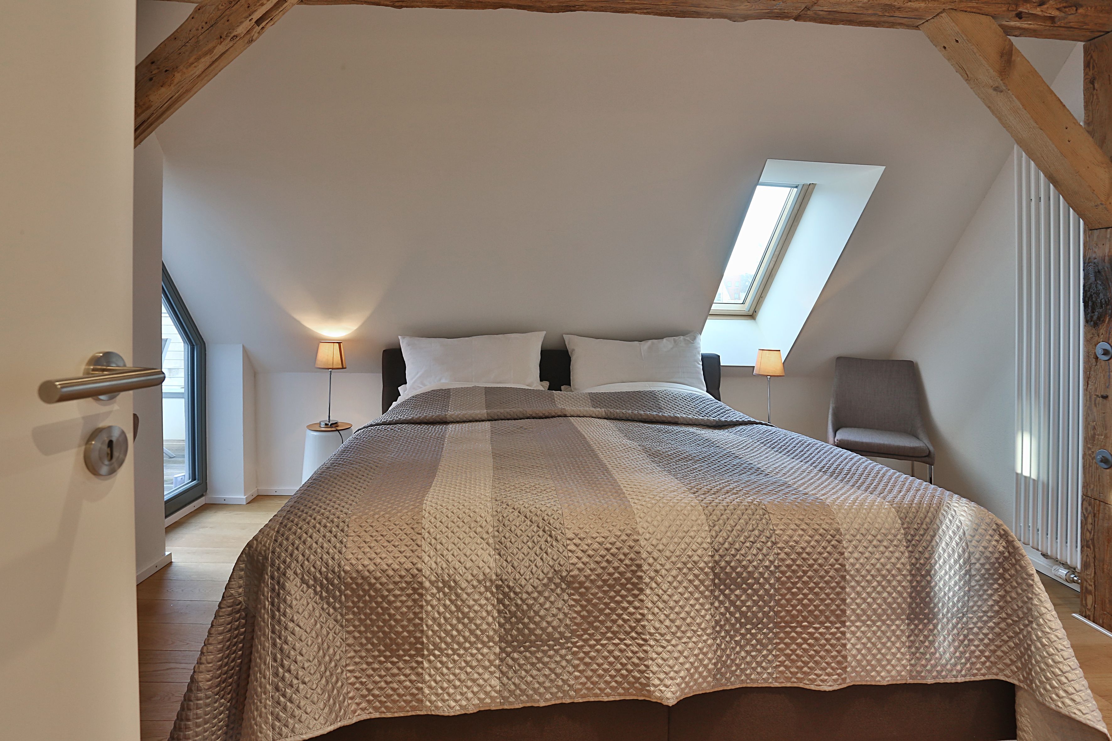 Une chambre avec un plafond en pente, un grand lit, deux lampes de chevet, une chaise et une fenêtre.