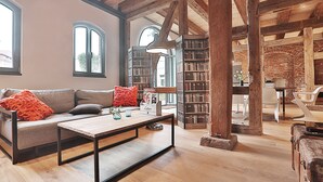A spacious living room with a wooden ceiling, a sofa with orange cushions, a coffee table, and a bookshelf filled with books. - Backsteinspeicher Stralsund (Stralsund)
